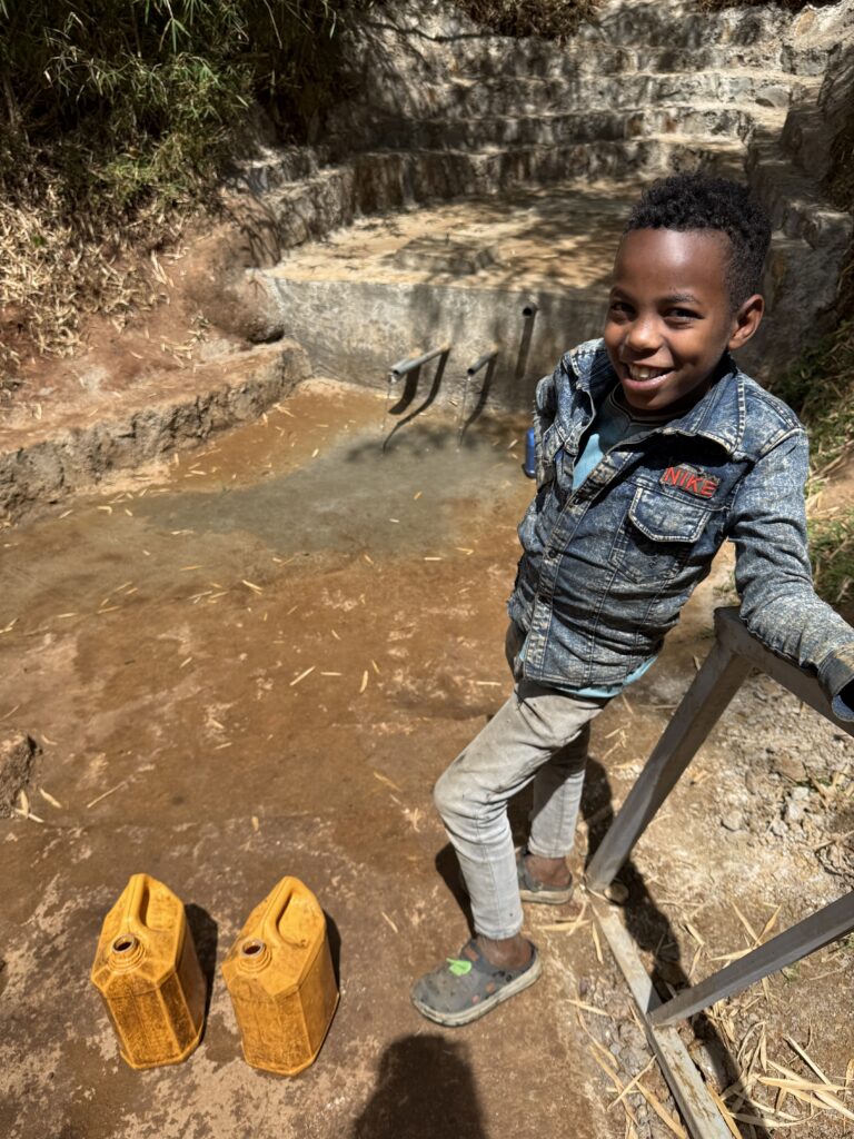 A young boy standing before an Hawassa Hope clean water site with water jugs in the picture. He has a big smile on his face. 
